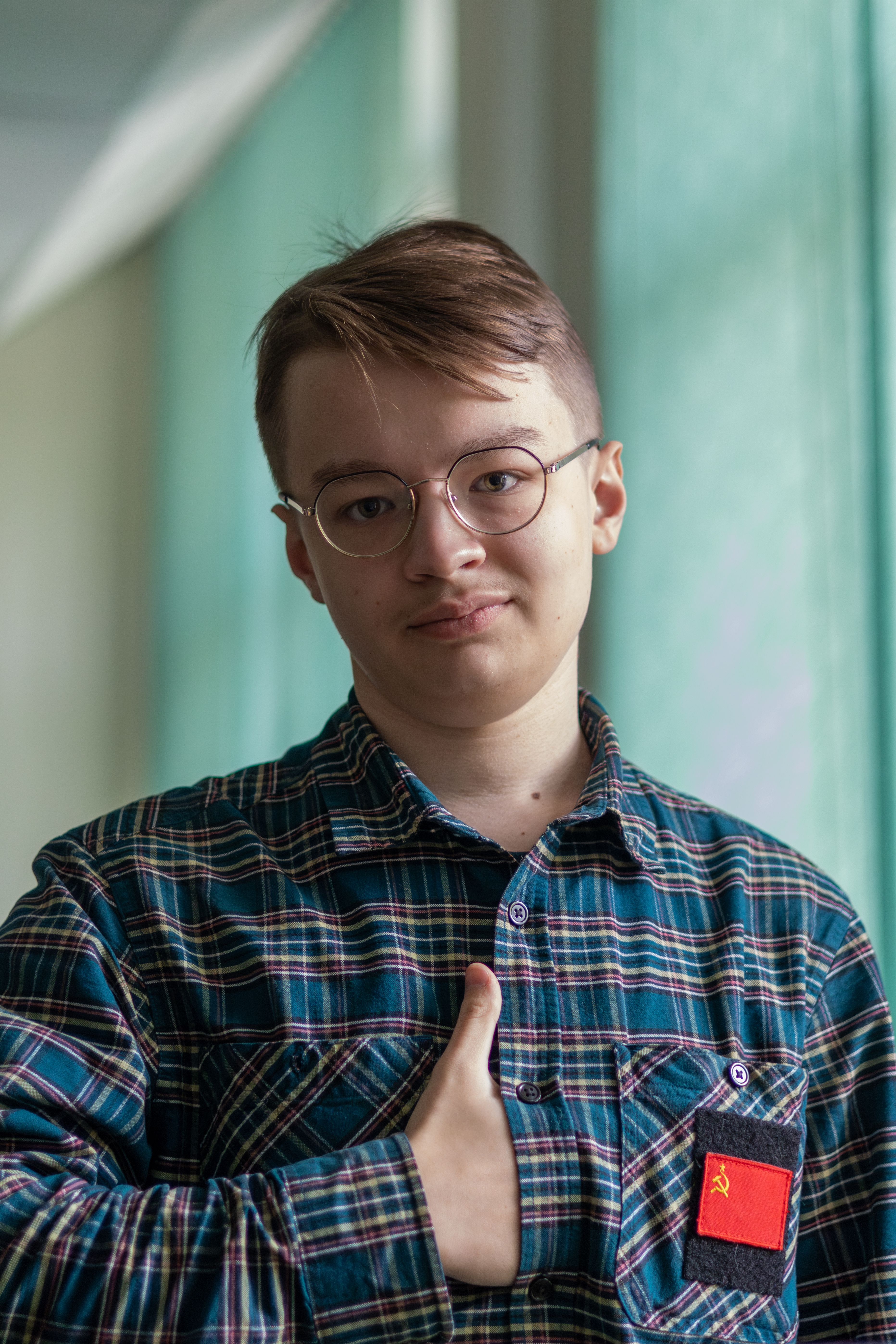 A young man wearing glasses and a plaid shirt with a red patch indoors, expressing confidence.