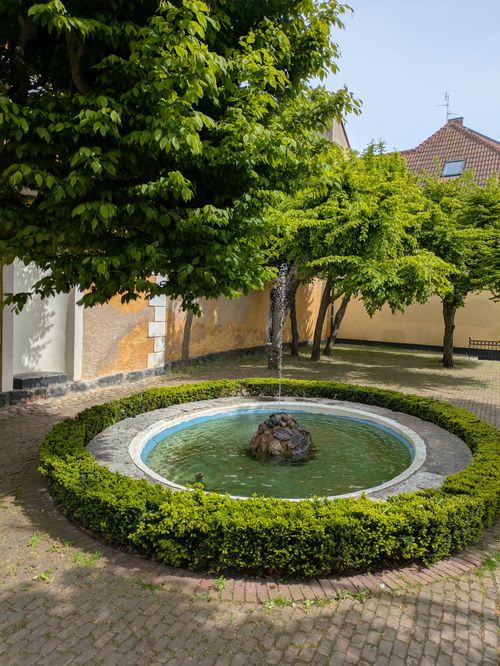Free A peaceful courtyard featuring a fountain surrounded by trees and a hedge, perfect for outdoor relaxation. Stock Photo