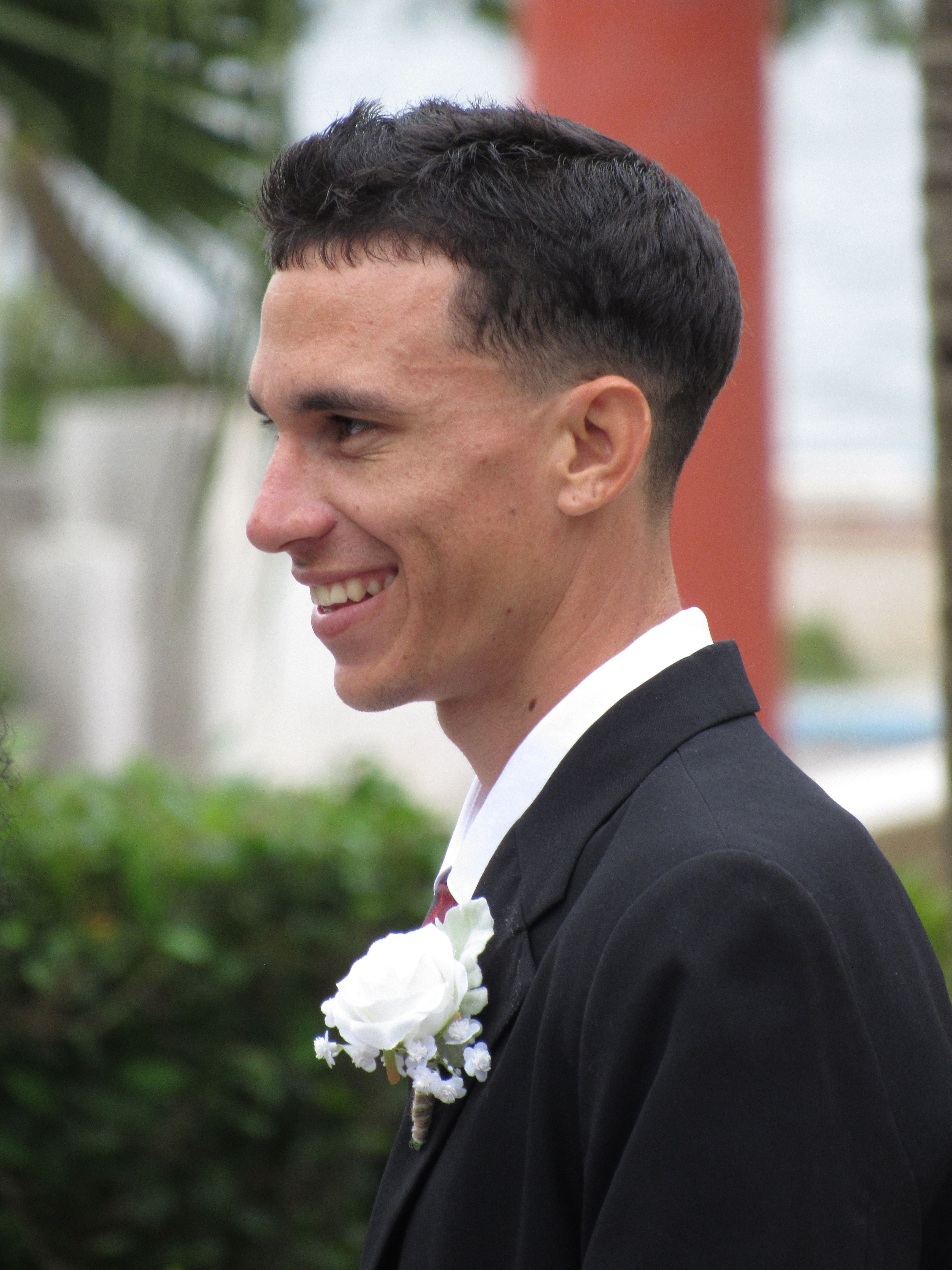 A smiling man in a suit with a white boutonniere in an outdoor setting.
