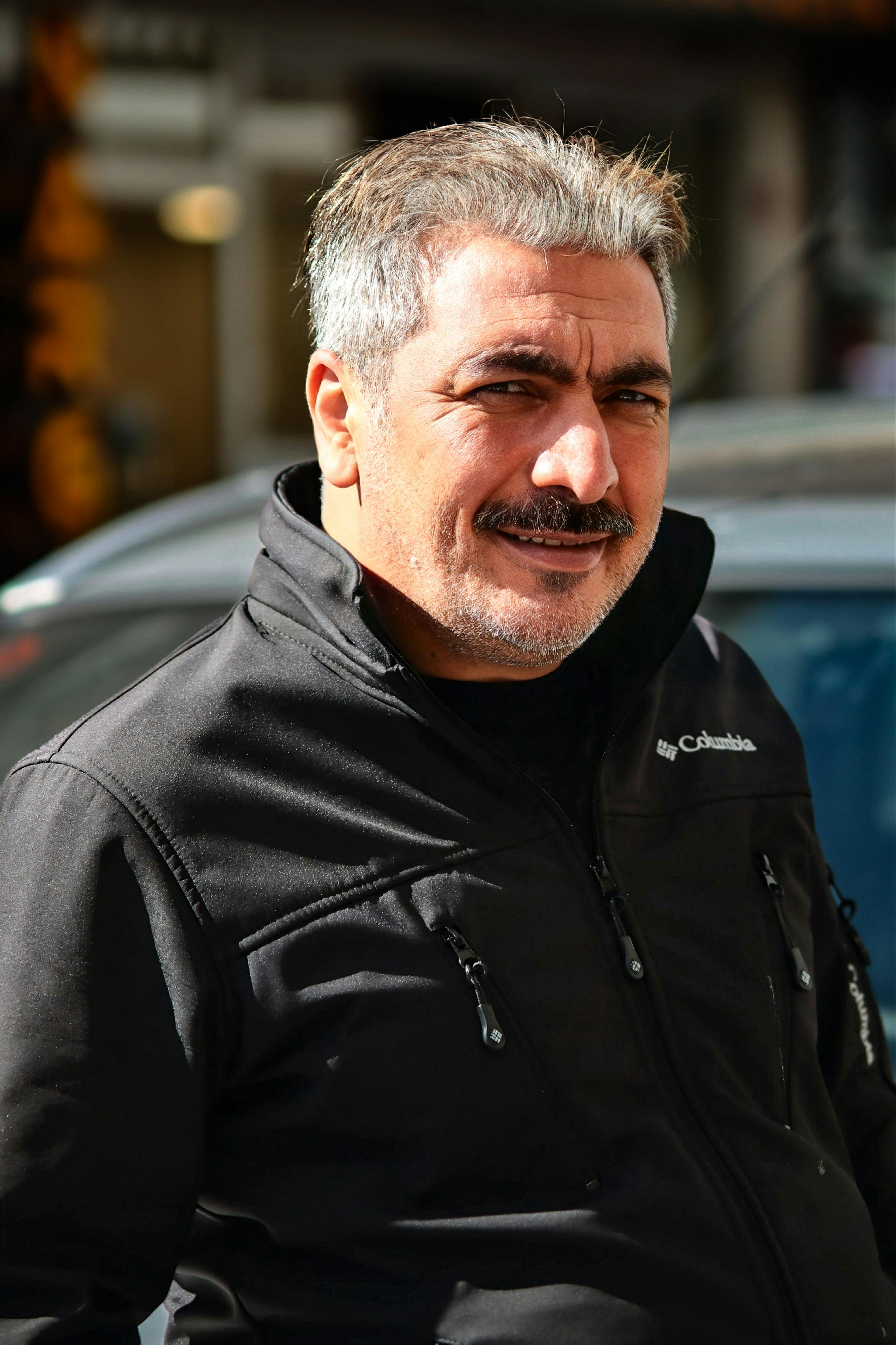 Smiling mature man posing outdoors in a black jacket, captured in natural daylight.