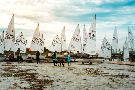 Group of people with sailboats on sandy beach, colorful sunset sky, leisure activity.