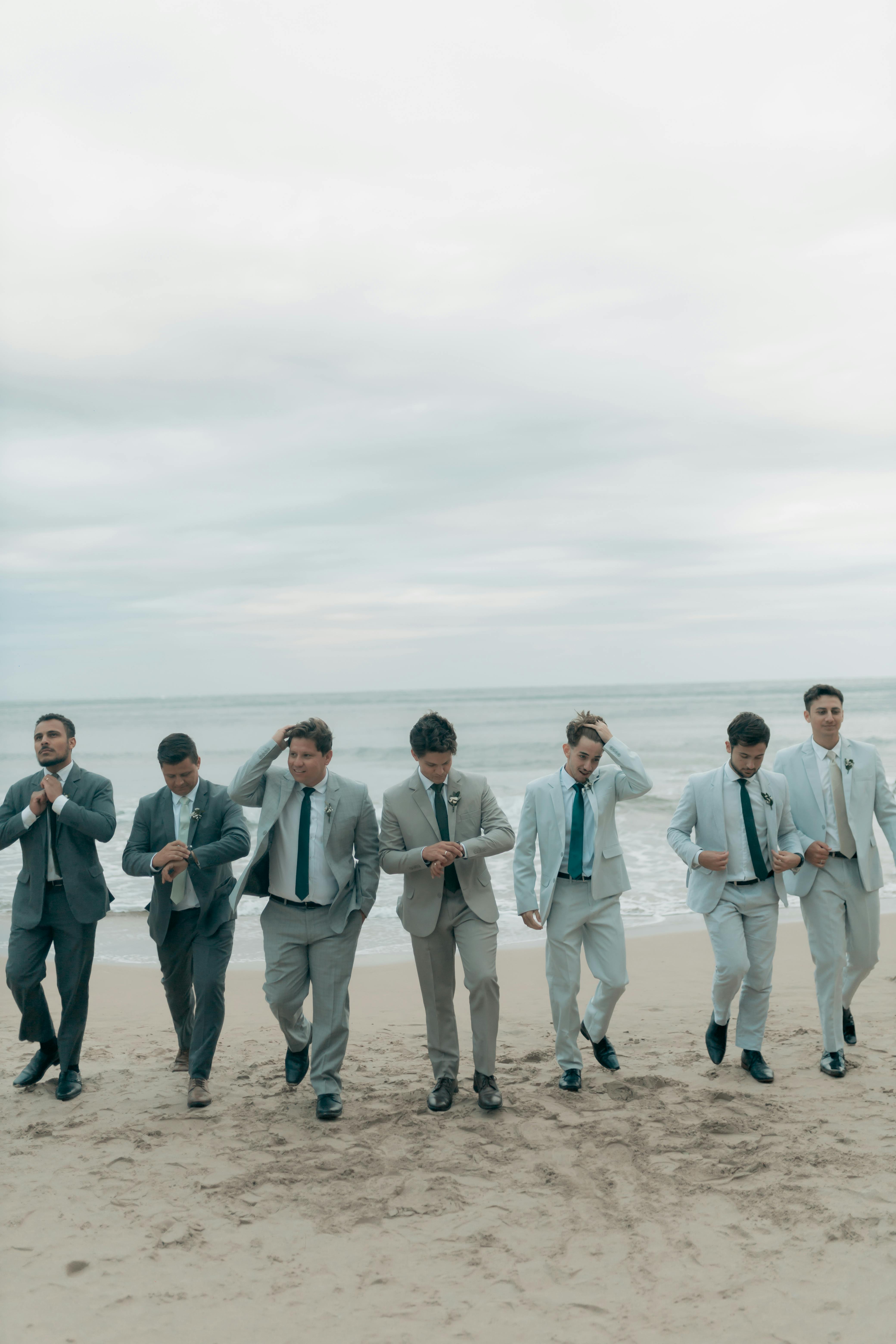 Seven groomsmen in suits walking along a beach, showcasing stylish wedding attire.