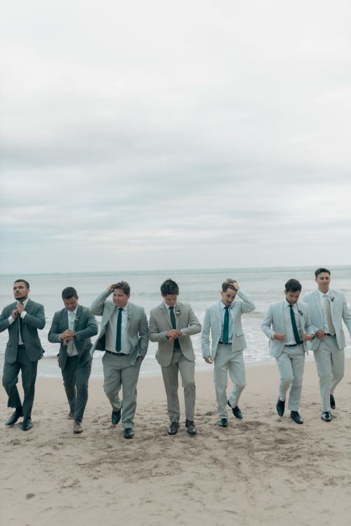 Free Seven groomsmen in suits walking along a beach, showcasing stylish wedding attire. Stock Photo