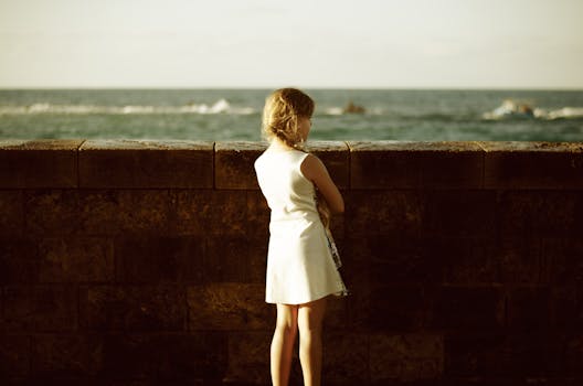 A young girl stands by a stone wall, gazing wistfully at the ocean under a warm sunset glow.