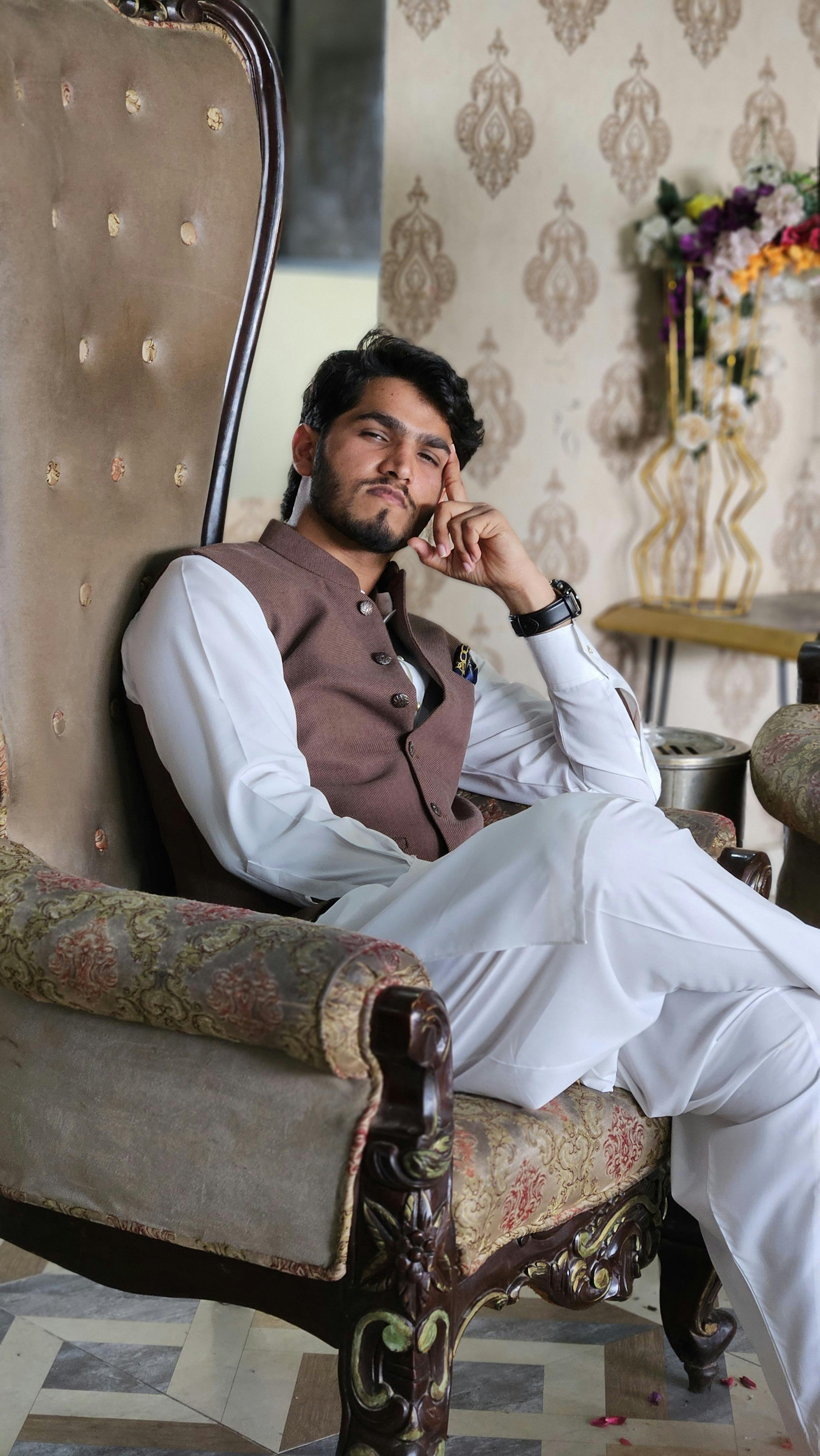 A stylish adult man wearing a waistcoat and shalwar kameez relaxing in an ornate chair indoors.