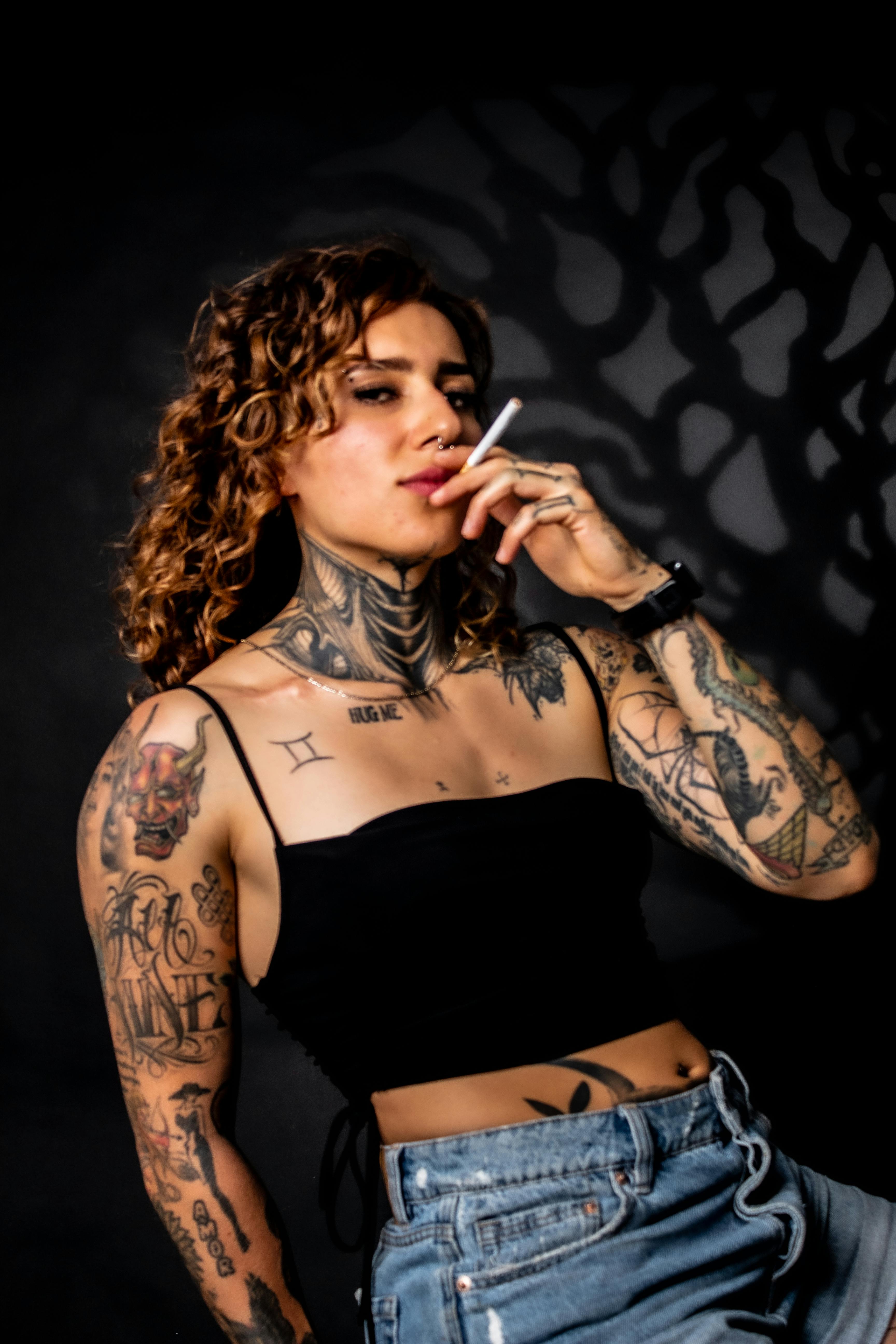 Portrait of a tattooed woman wearing a black top, smoking, against a dark backdrop.