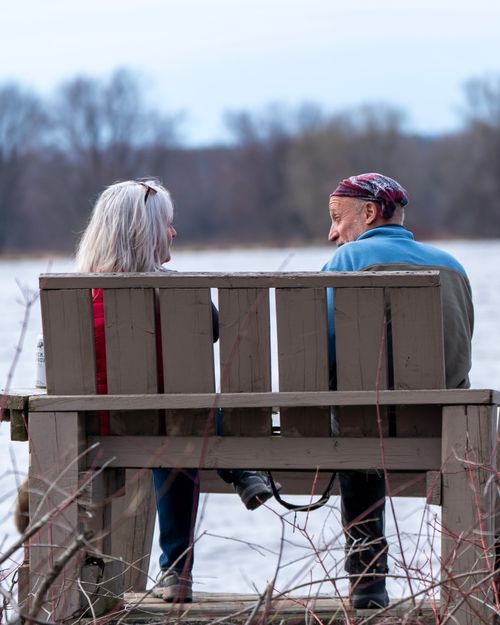 Free An elderly couple sits on a bench by a lake, enjoying nature in winter. Stock Photo