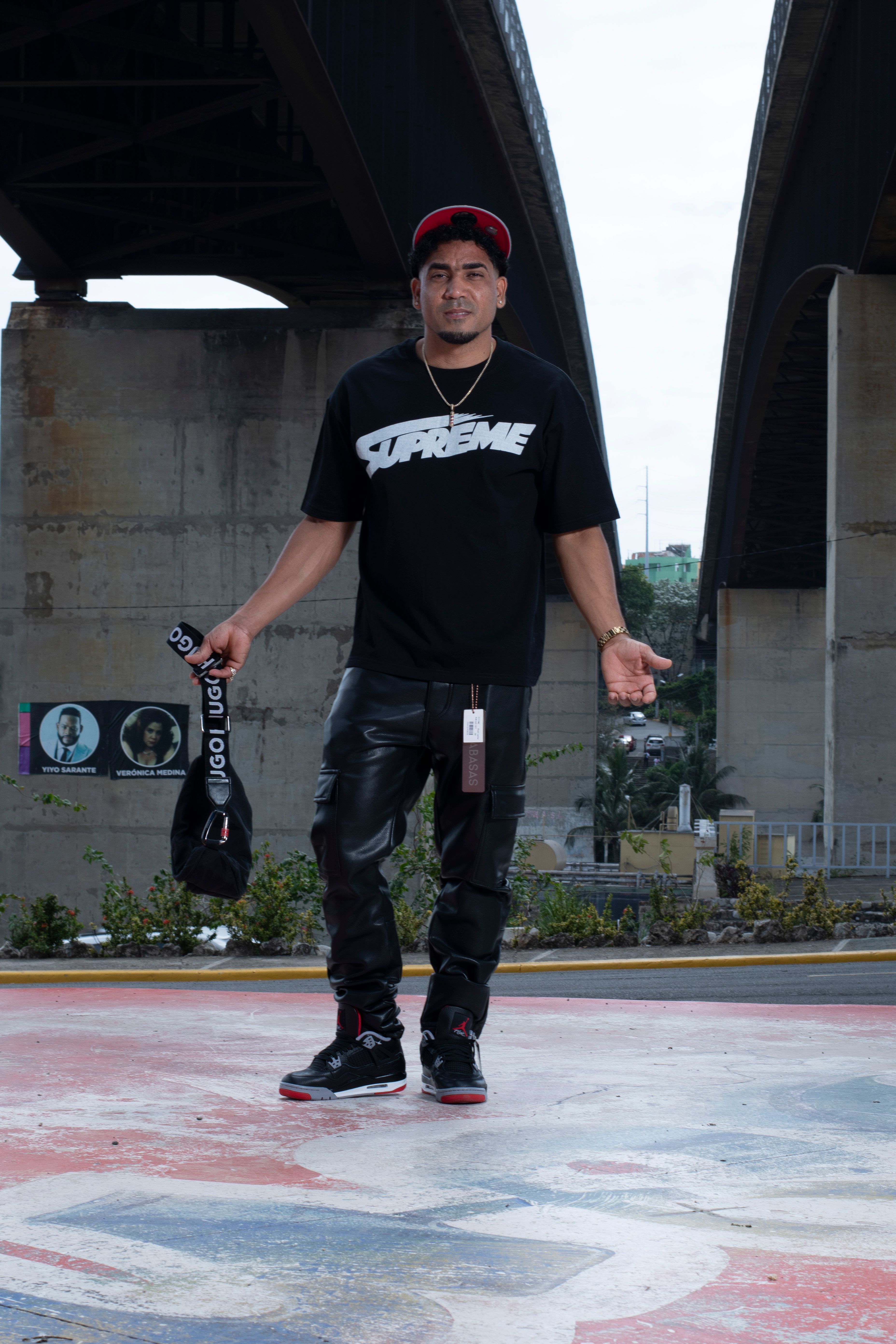 Man poses in urban street style with graphics under bridge, daytime.