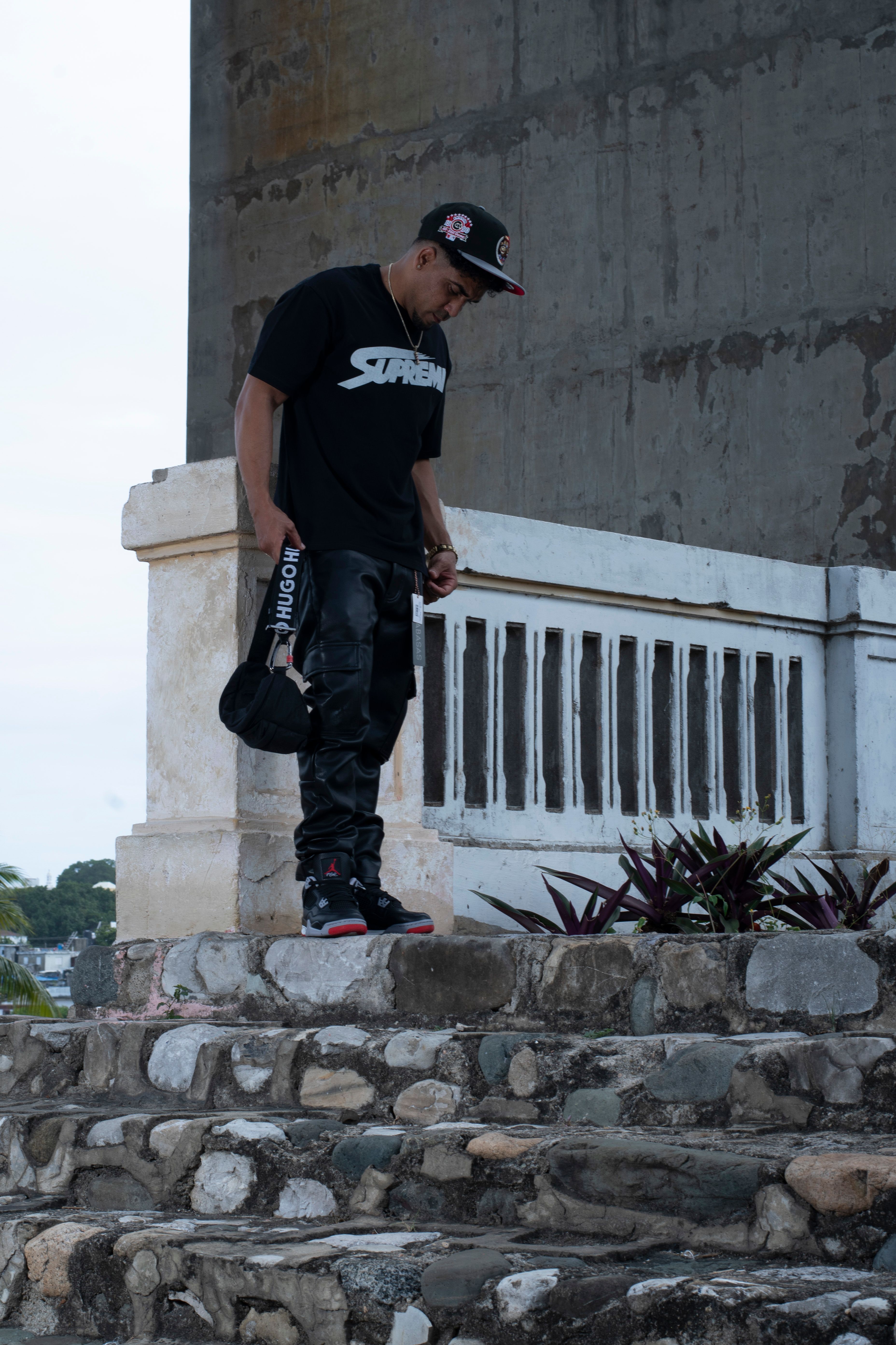 Young man in urban streetwear posing on stone steps beneath a concrete bridge.