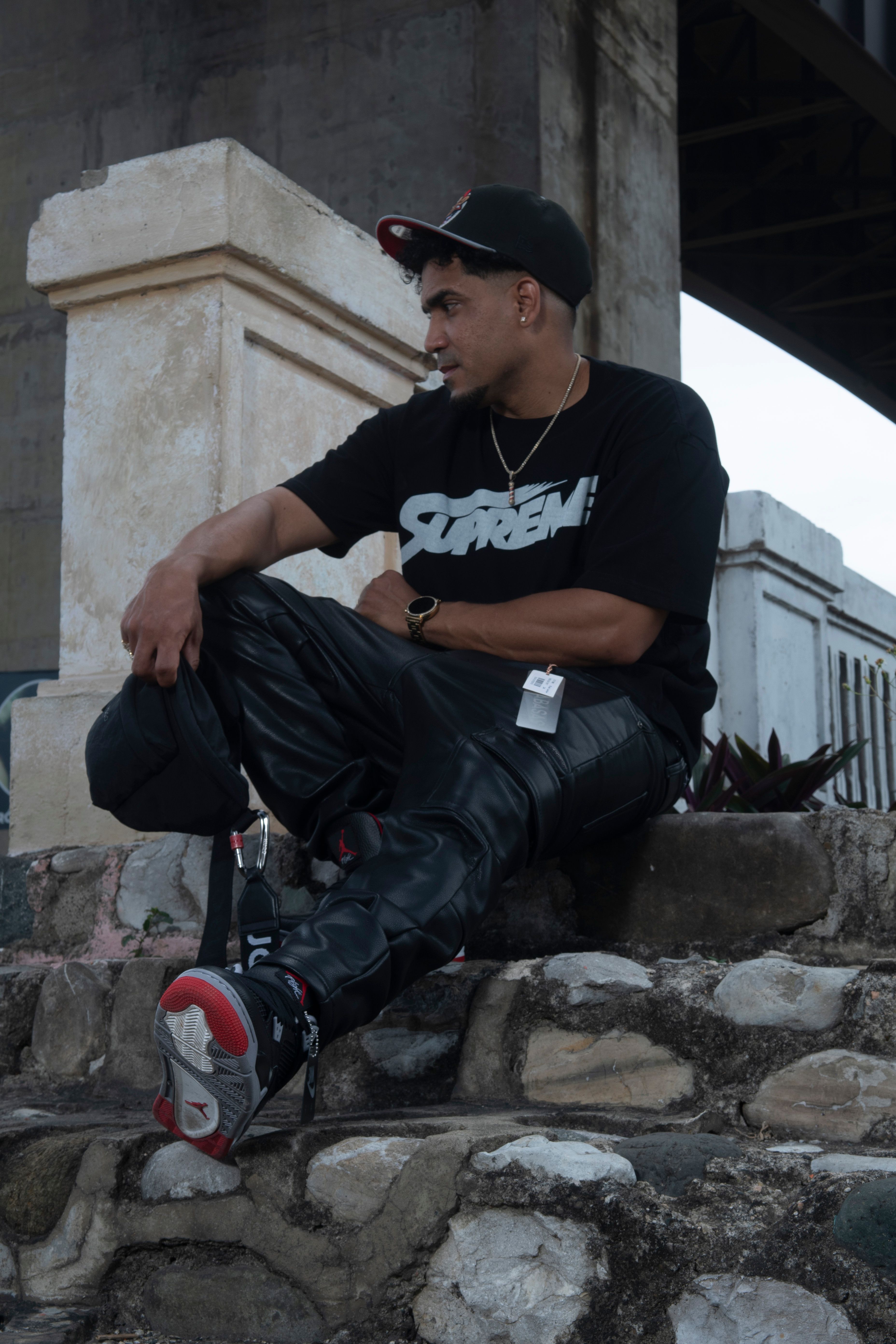 A stylish man in streetwear fashion sitting outdoors under a bridge.
