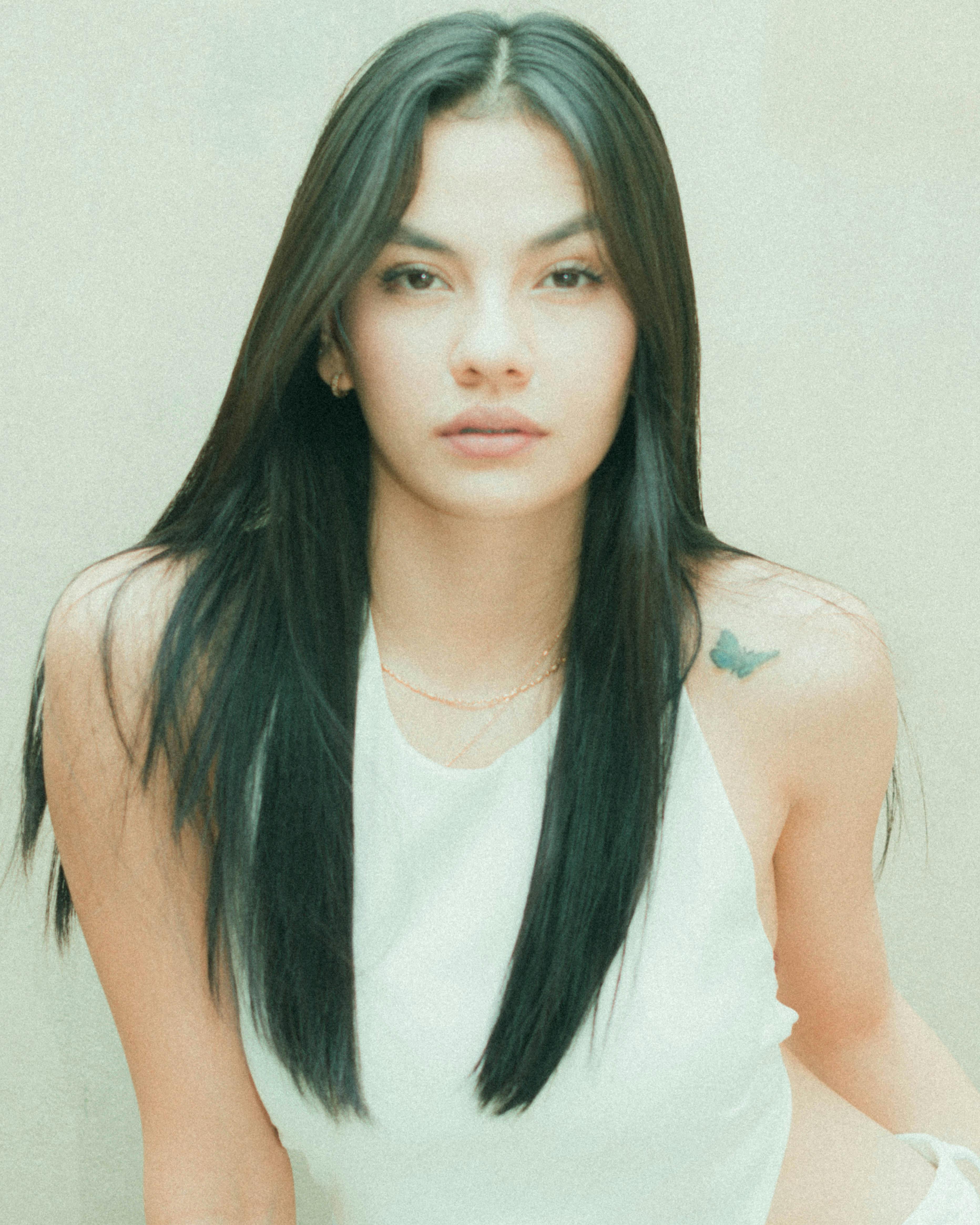 A portrait of a young woman with long hair and a tattoo, captured in natural light with a soft background.