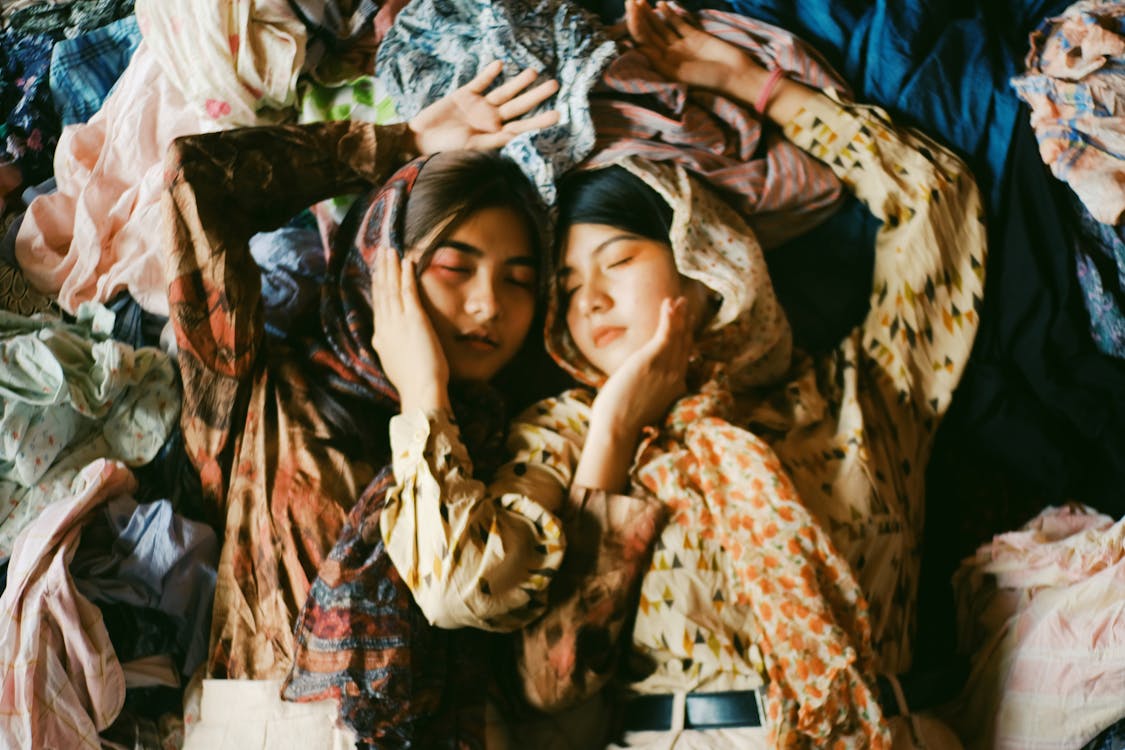 Two Women Wearing Earth-toned Printed Tops And Scarves Lying On A Pile Of Clothes