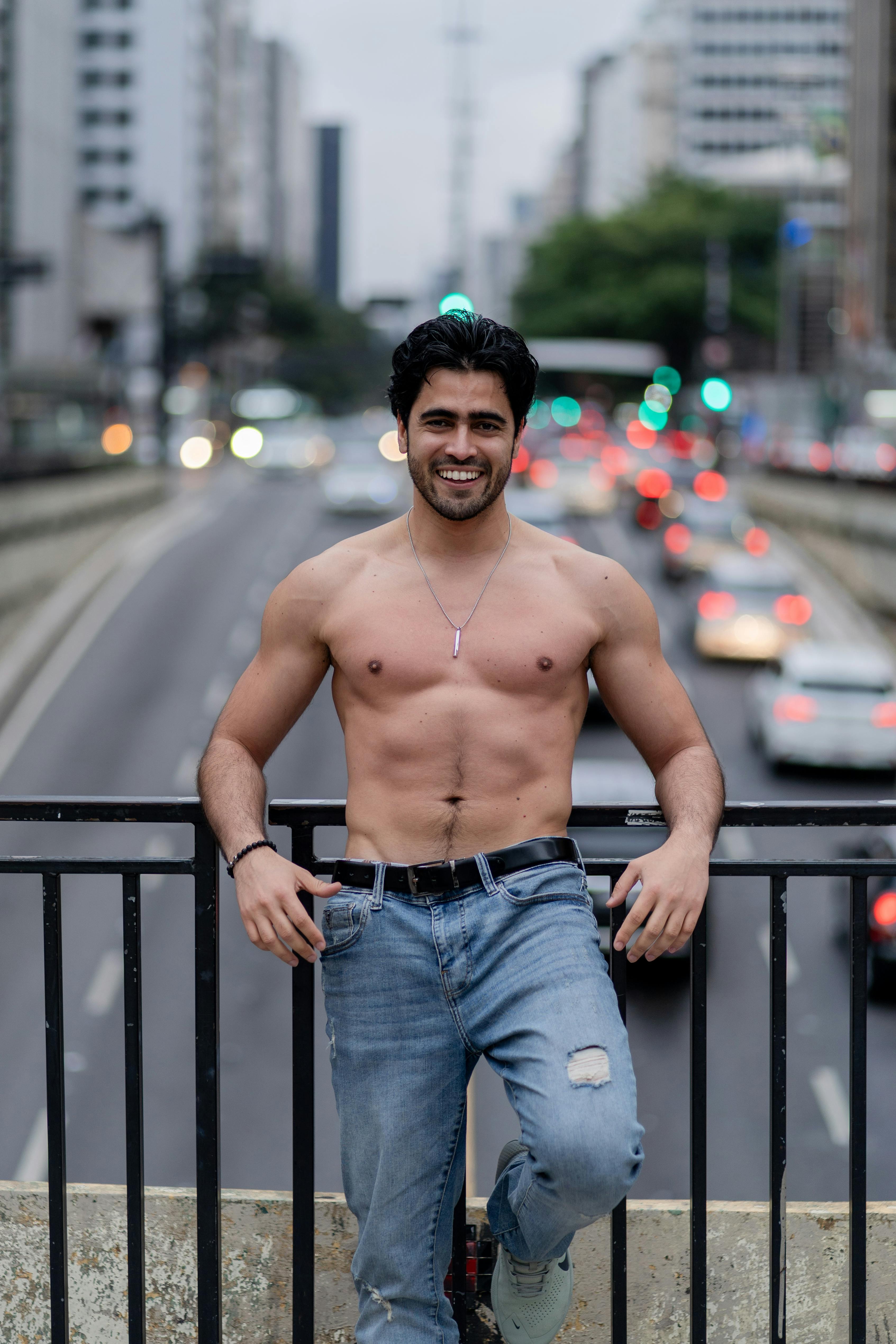 Shirtless man smiling on a city bridge during daytime traffic.