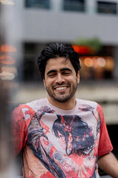 A cheerful man in an anime t-shirt enjoying a day outdoors with city lights in the background.