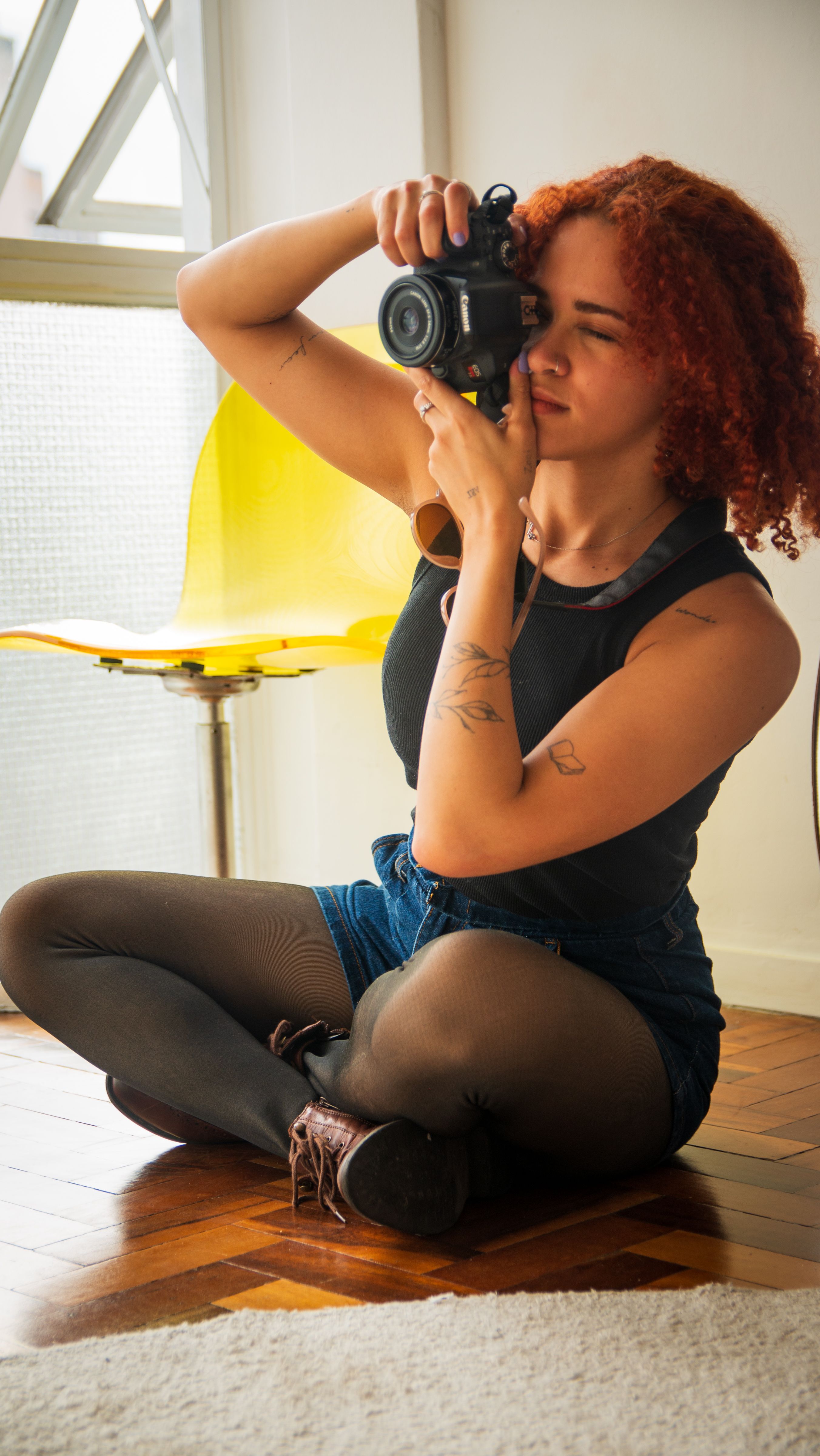 Young woman photographer sitting cross-legged indoors holding a camera, focused on capturing moments.