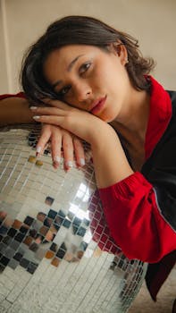Portrait of a woman in red jacket leaning on a reflective disco ball, indoors.
