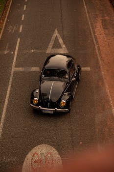 Aerial view of a classic black Volkswagen Beetle driving on a city road with autumn leaves.