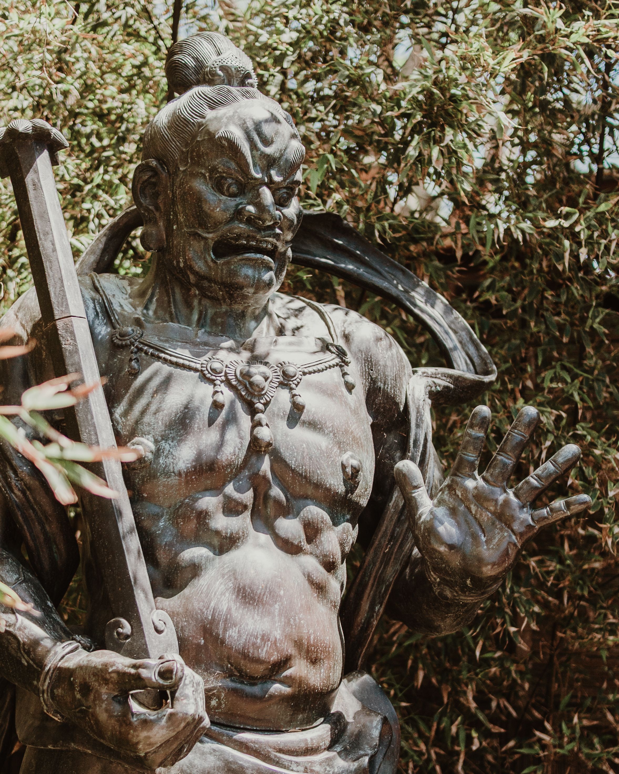 Bronze warrior statue with fierce expression surrounded by lush greenery.
