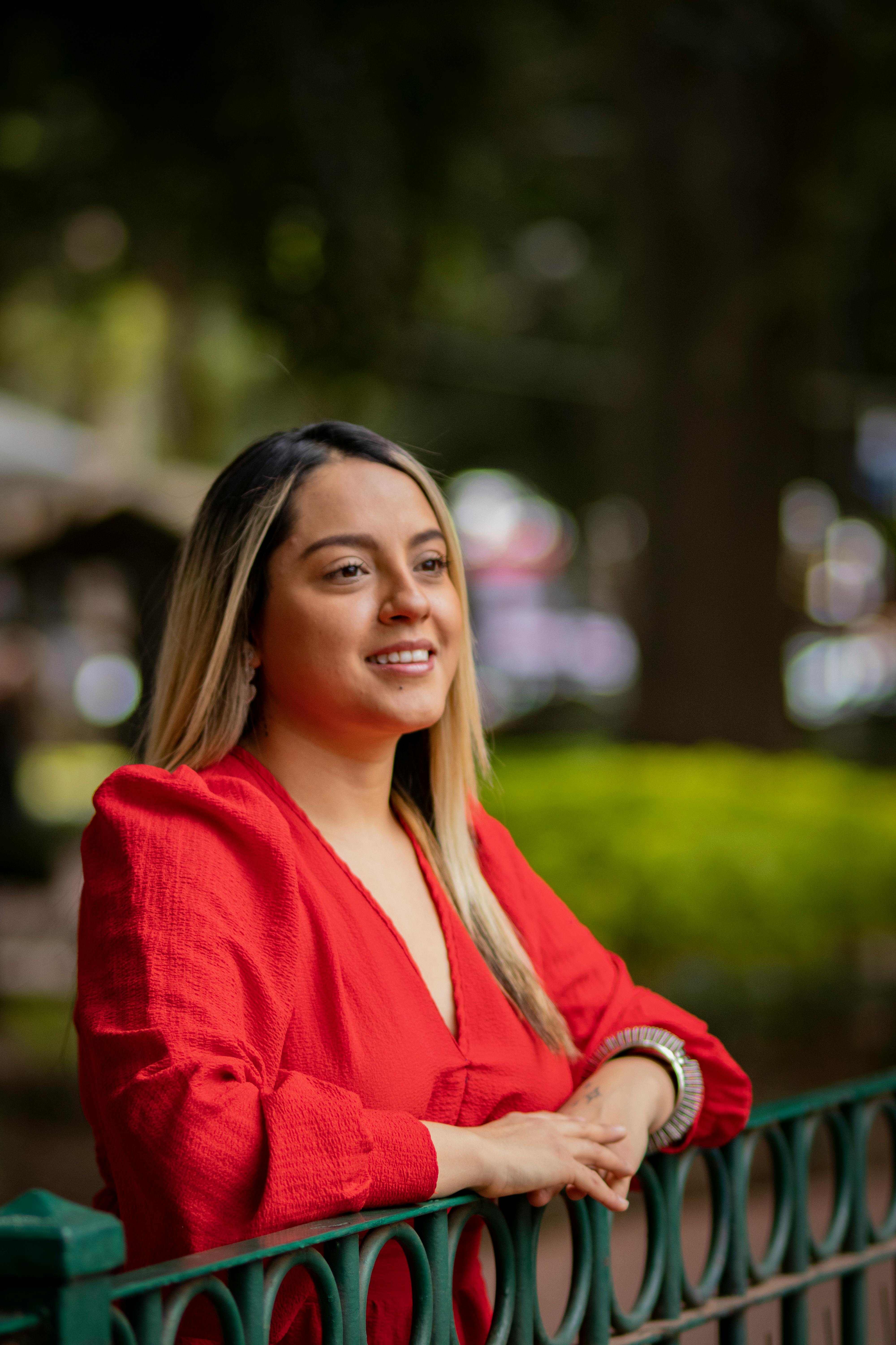 Portrait of a smiling woman leaning on a fence wearing a red dress in a park.