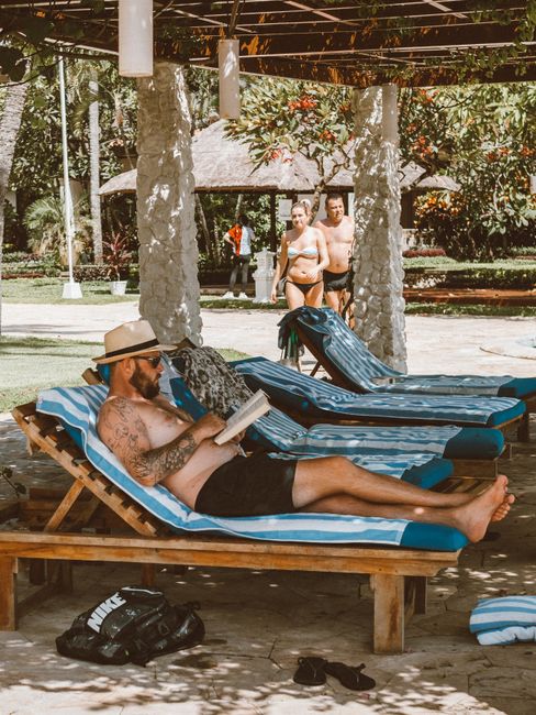 Man reading by the poolside with others in the background, enjoying a sunny day.