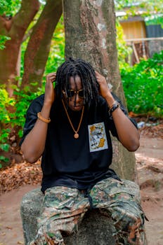 Young man in casual streetwear sitting outdoors on a stone, surrounded by greenery.
