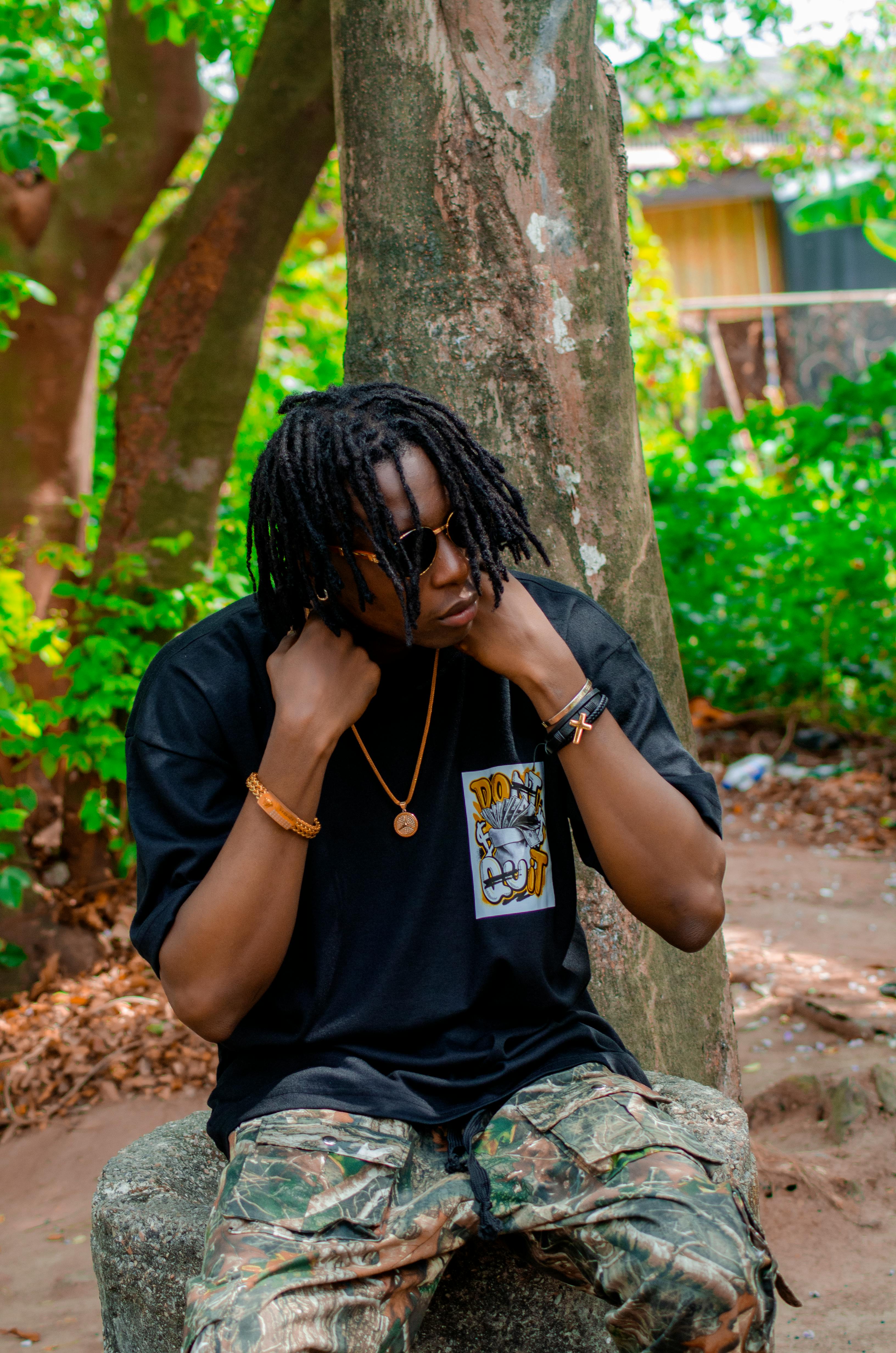 A man in casual attire with sunglasses sitting outdoors by a tree, exuding cool vibes.