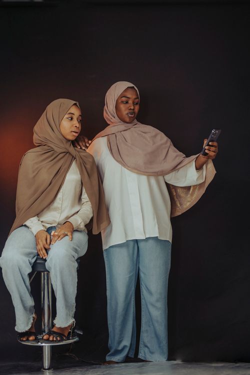 Free Two women wearing hijabs take a selfie against a dark backdrop for a portrait session. Stock Photo