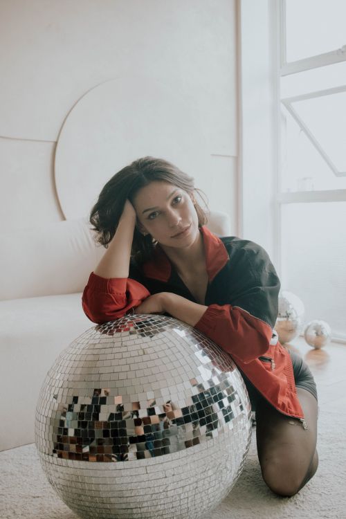 Free A woman in a red jacket rests on a large disco ball in a bright, cozy indoor space. Stock Photo