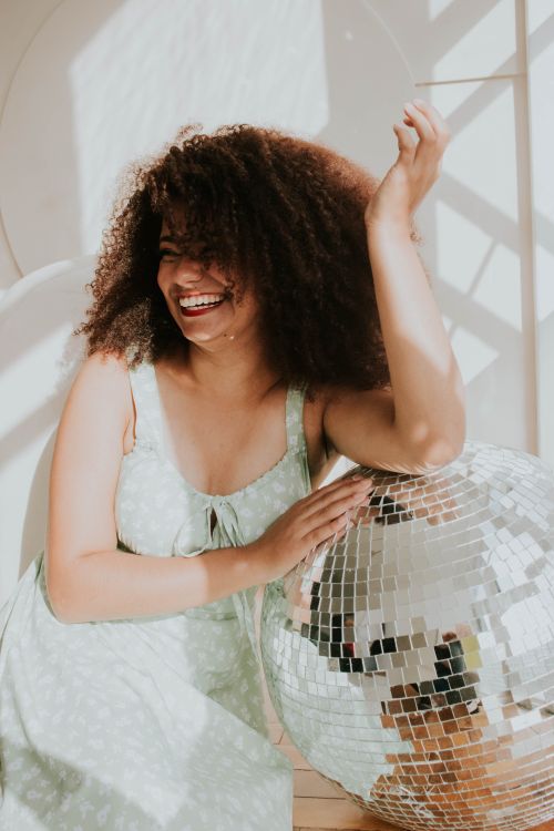Free Joyful young woman with curly hair smiling and sitting beside a sparkling disco ball in bright sunlight. Stock Photo