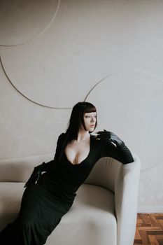 Stylish woman wearing black gown poses gracefully on a contemporary white sofa, exuding elegance.