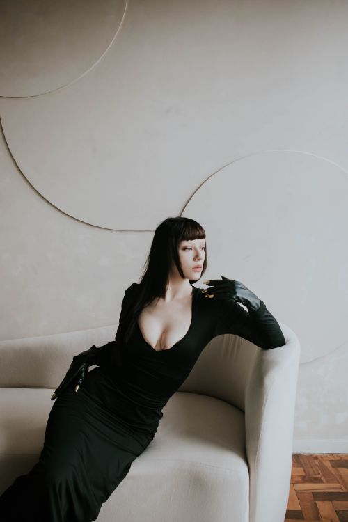 Free Stylish woman wearing black gown poses gracefully on a contemporary white sofa, exuding elegance. Stock Photo
