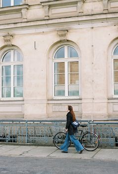 A woman walks beside her bike past a historic building facade.