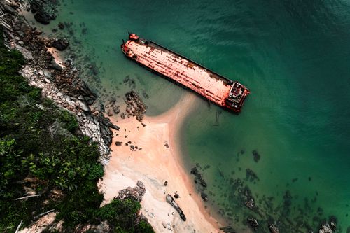 Free Aerial view of a rusted shipwreck on a remote sandy beach surrounded by rocky cliffs and green water. Stock Photo