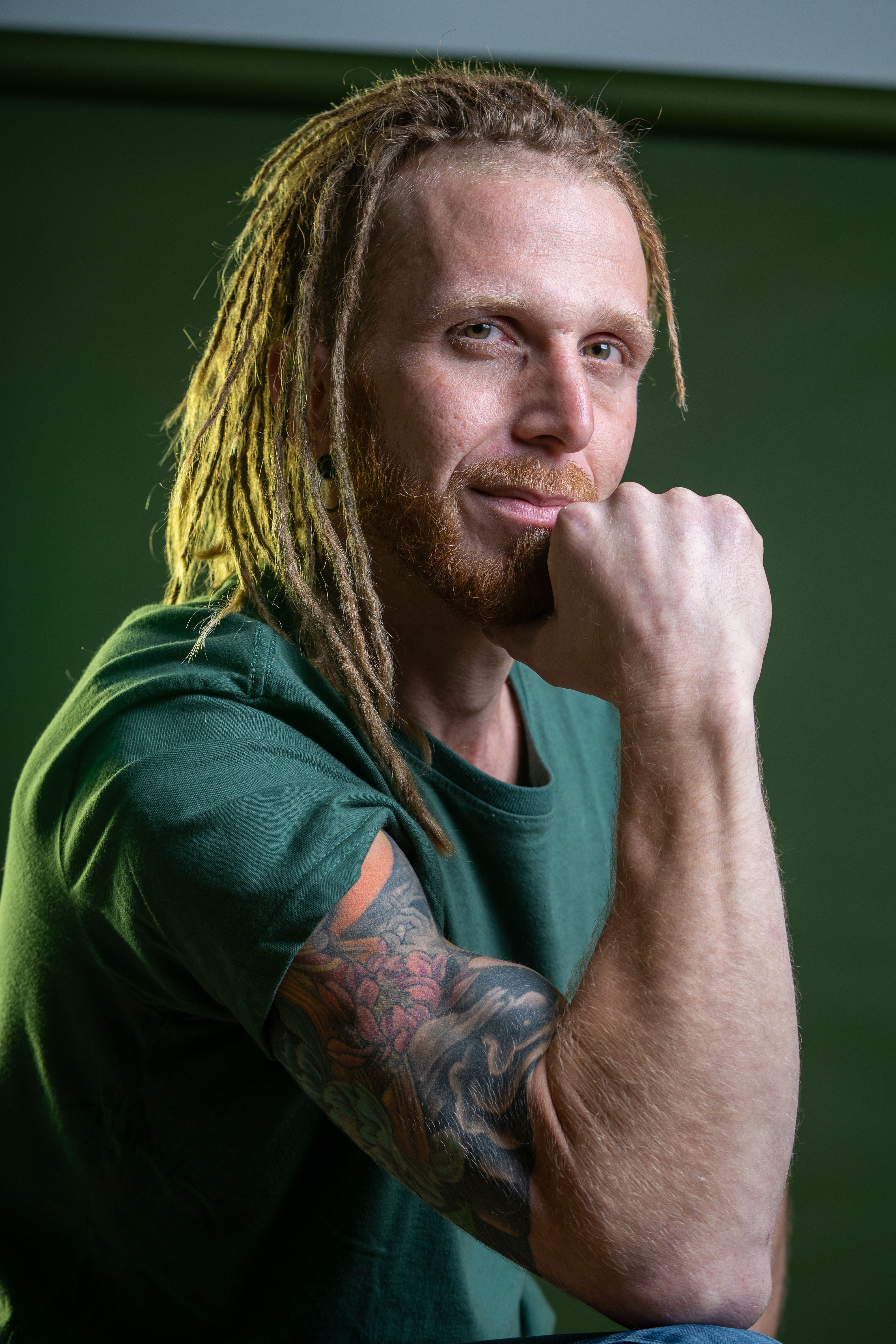 Close-up portrait of a man with dreadlocks and tattoo sleeve, showcasing his thoughtful pose against a green background.