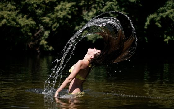 Graceful woman in yellow bikini flips hair in a refreshing summer lake, capturing vibrant energy and motion.