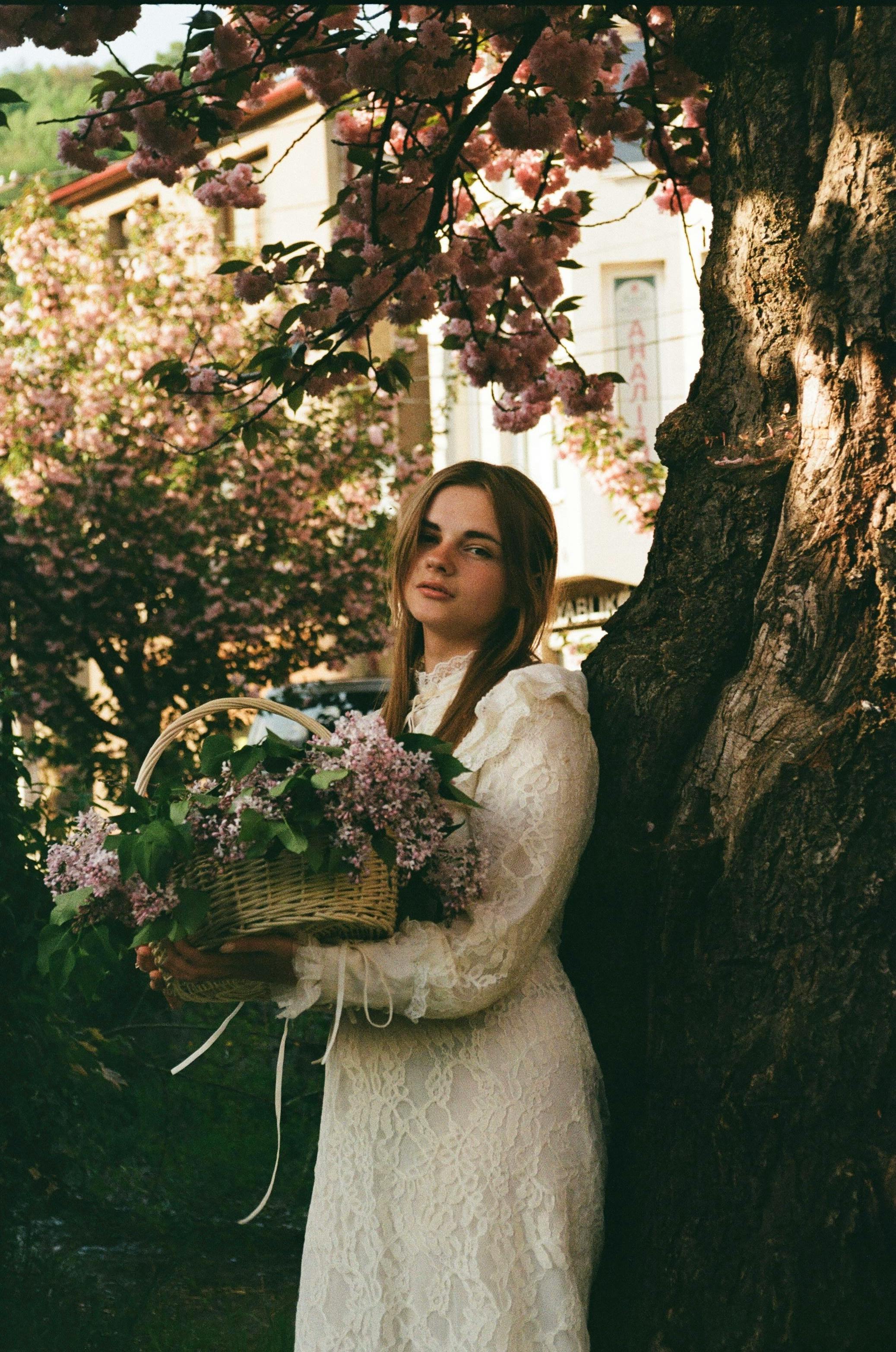 A woman in a lace dress holds a floral basket under blooming trees.