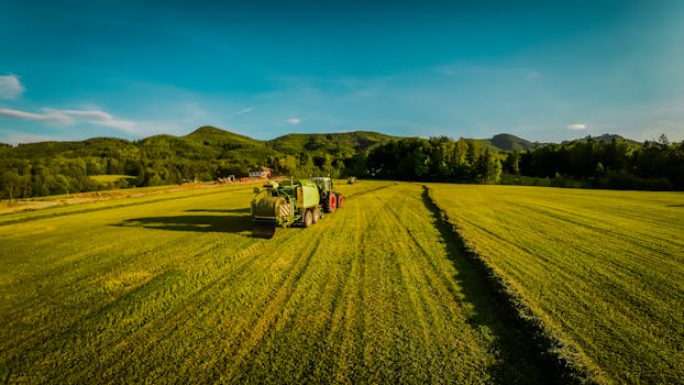 Aerial photo of tractors working on lush green farm fields in rural Austria with forest and hills in the background.