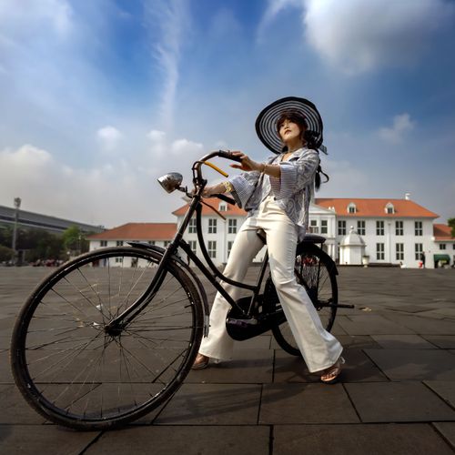 Free Woman in hat and white outfit poses on bicycle in front of historical building under blue sky. Stock Photo