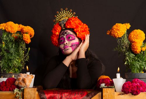Free Adult woman with traditional Dia de los Muertos makeup and marigold crown, sitting by decorated altar. Stock Photo