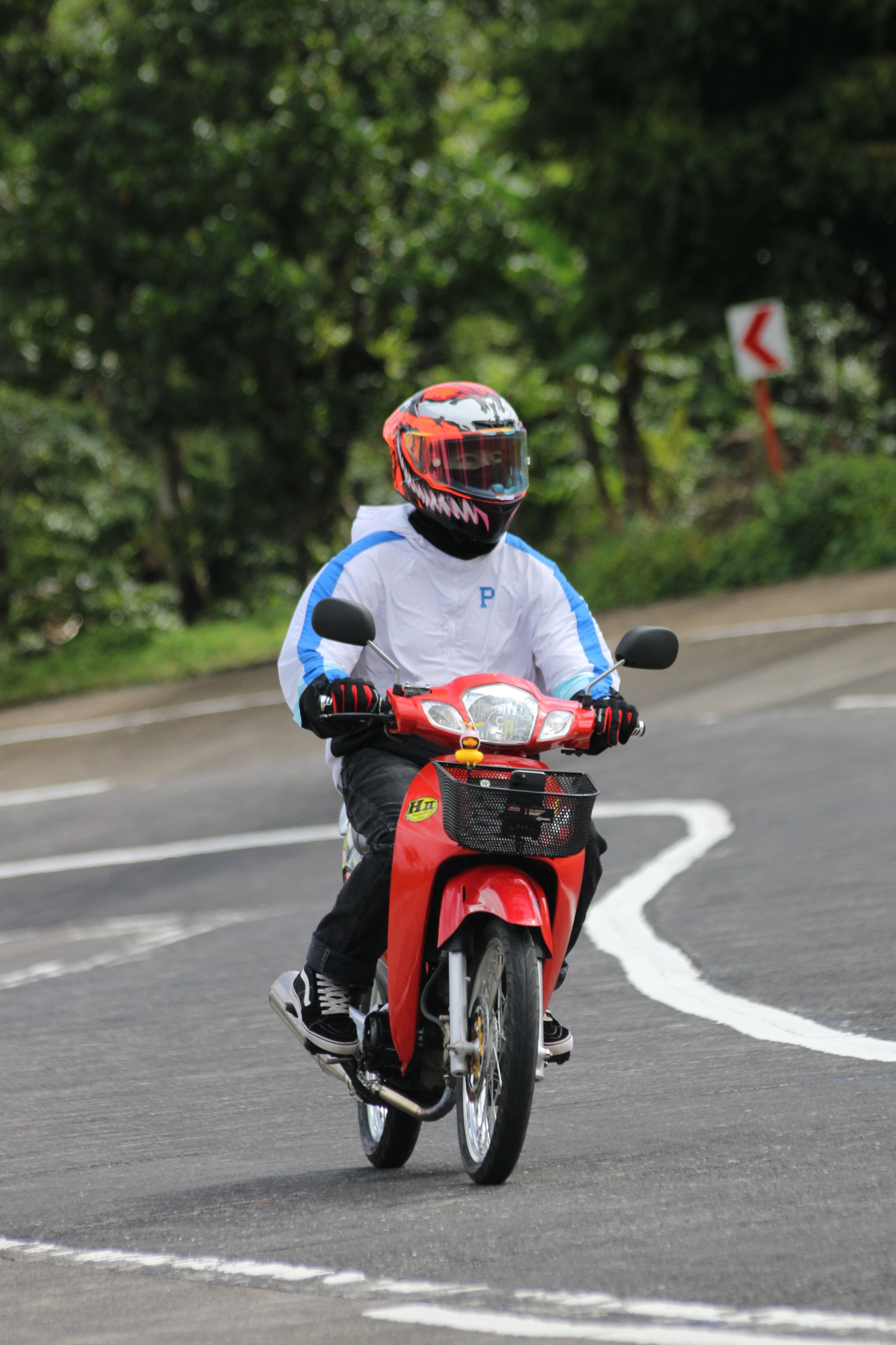 A motorcyclist rides a red scooter on a curved road, wearing a helmet and riding gear.