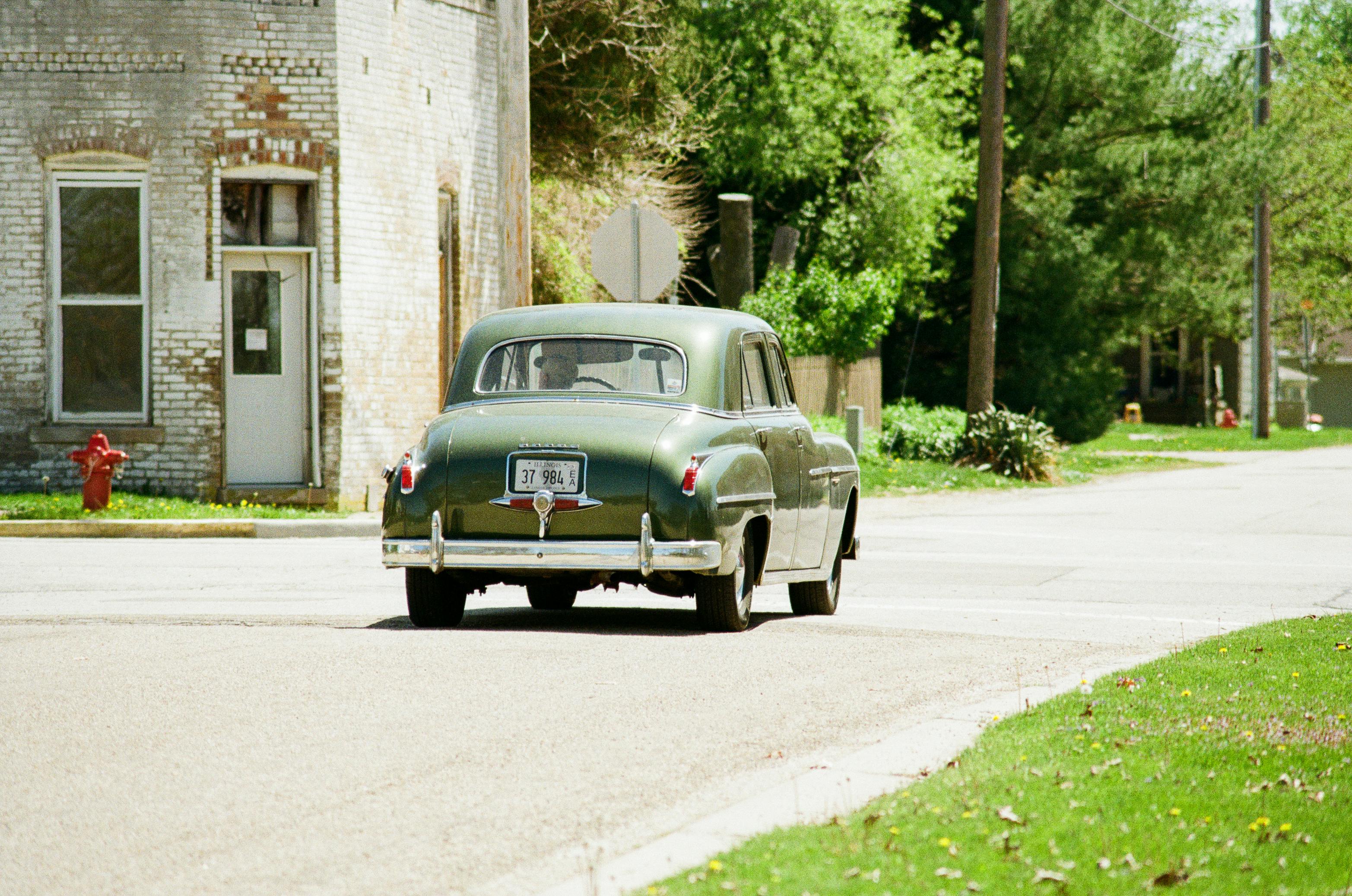 Green vintage car driving on a quiet neighborhood street next to an old building.