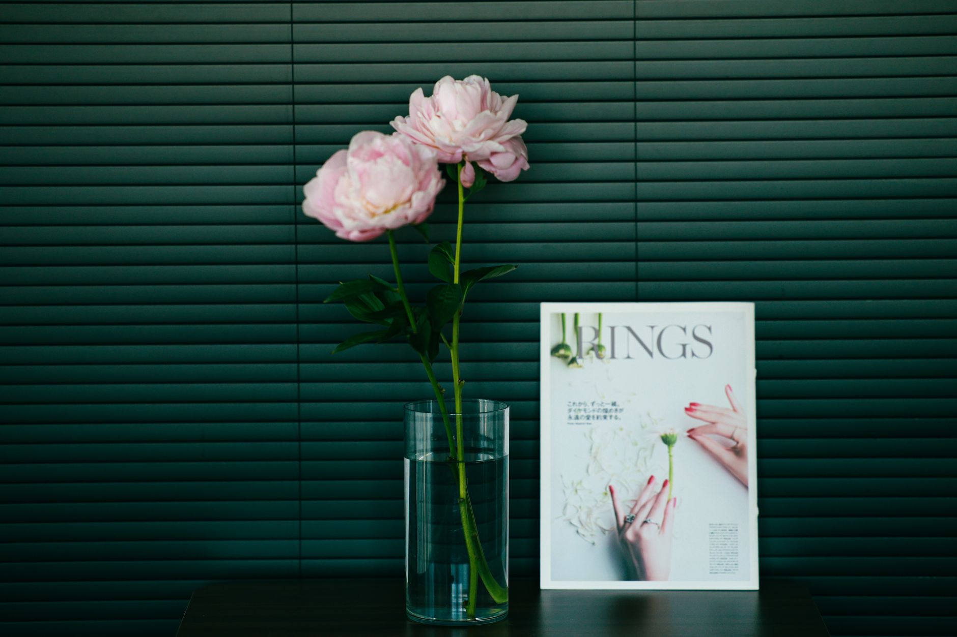 Elegant pink peonies in glass vase beside fashion magazine on dark background.