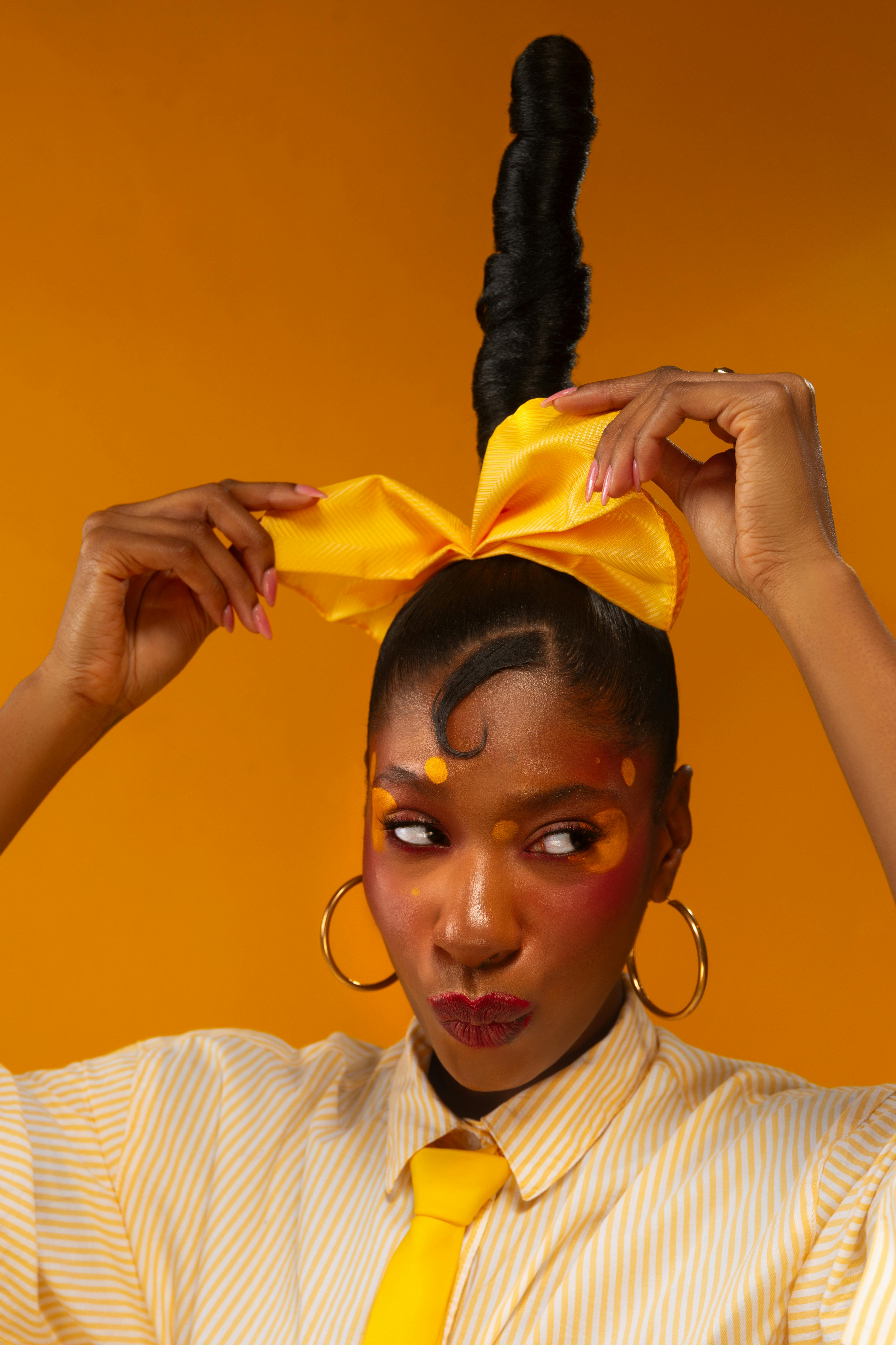 Fashionable woman poses with unique hairstyle and vivid makeup against a bright orange background.