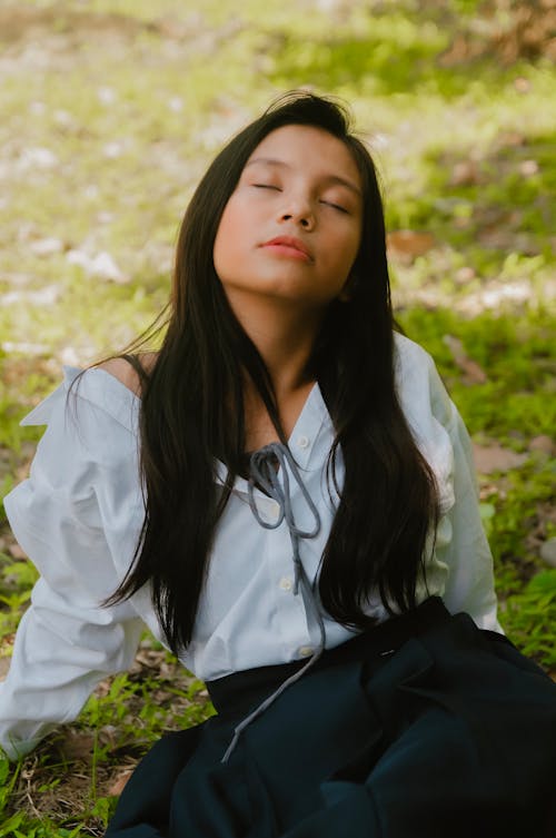 Selective Focus Photo of Woman Sitting on Grass Field Posing With Her Eyes Closed