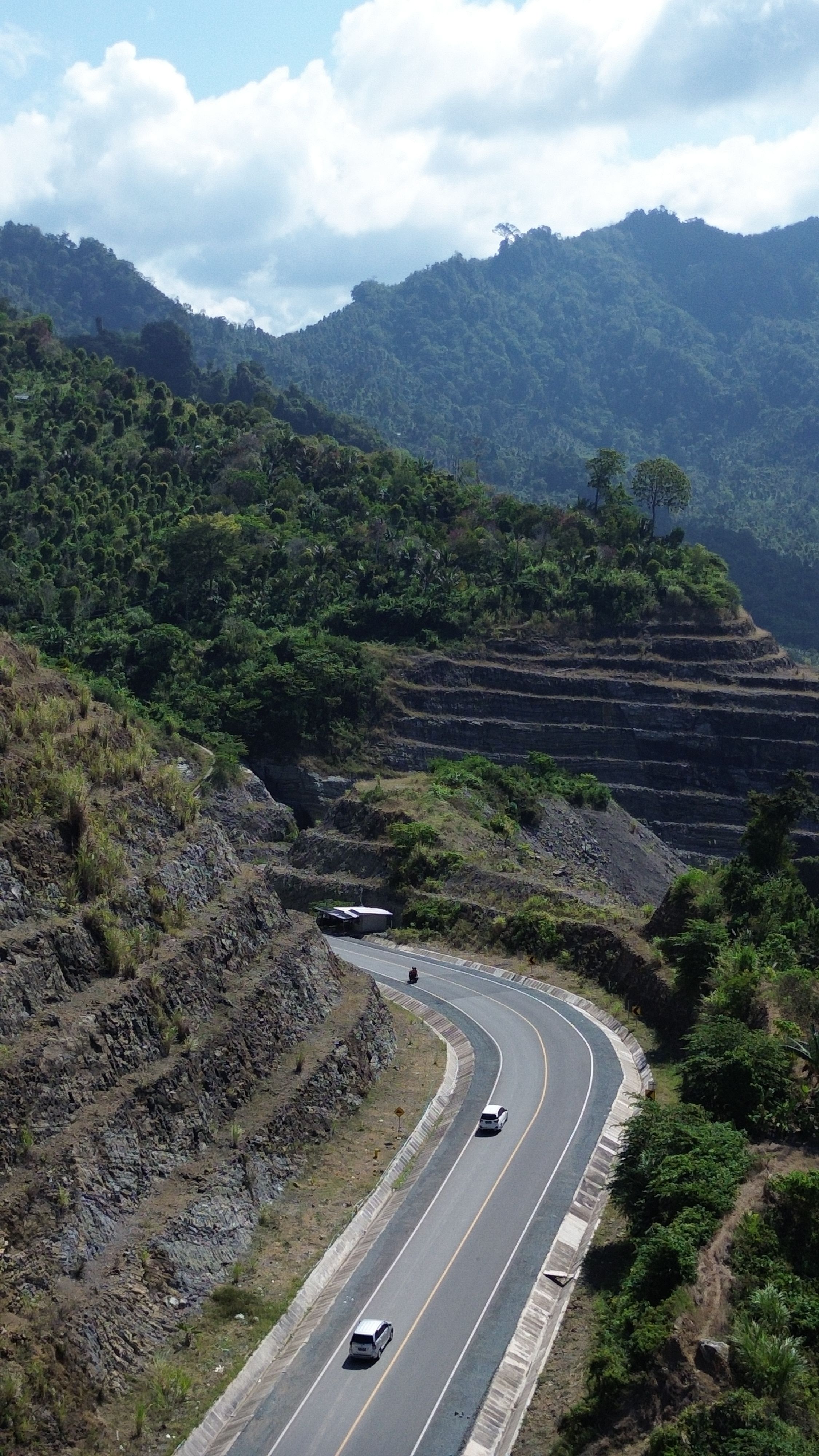 Aerial view of a winding road cutting through a lush, mountainous region in Indonesia, captured on a sunny day.