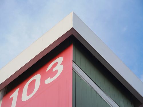 Free Close-up of a modern building corner with number 103 on a red panel against a blue sky. Stock Photo