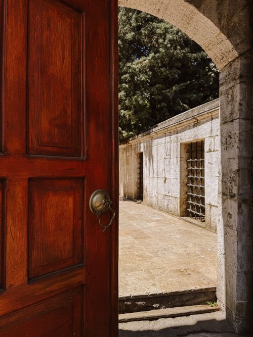 Free A red wooden door opens to reveal a sunlit stone courtyard with trees. Stock Photo