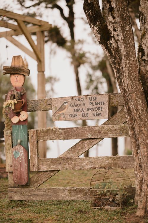 Free Rustic farm gate with carved wooden figure, birdcage, and motivation sign in countryside setting. Stock Photo