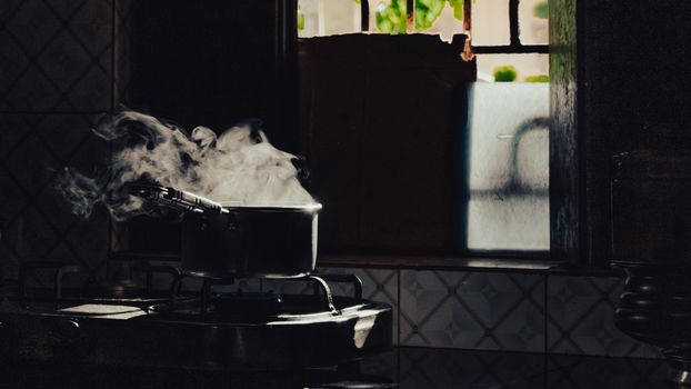 Atmospheric shot of steam rising from a pot on a stove in a dimly lit kitchen with window light.