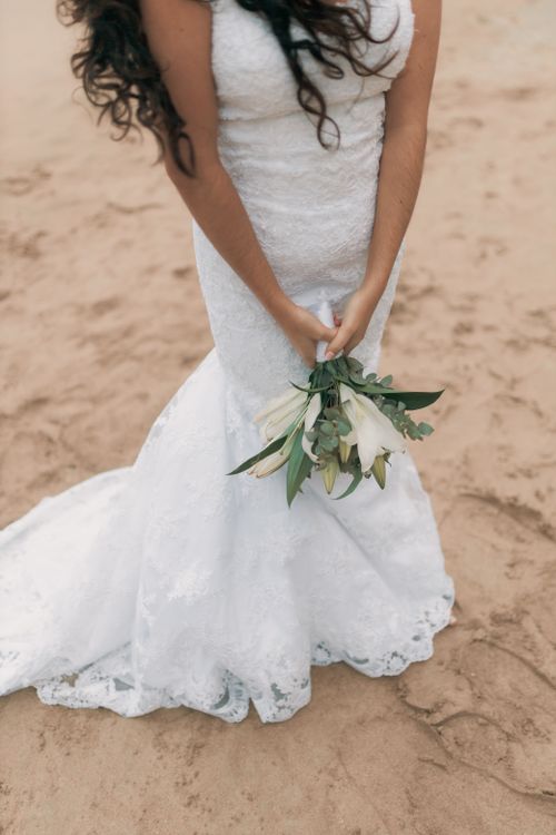 Free A bride in a white wedding gown holds a bouquet of white lilies on a sandy beach. Stock Photo